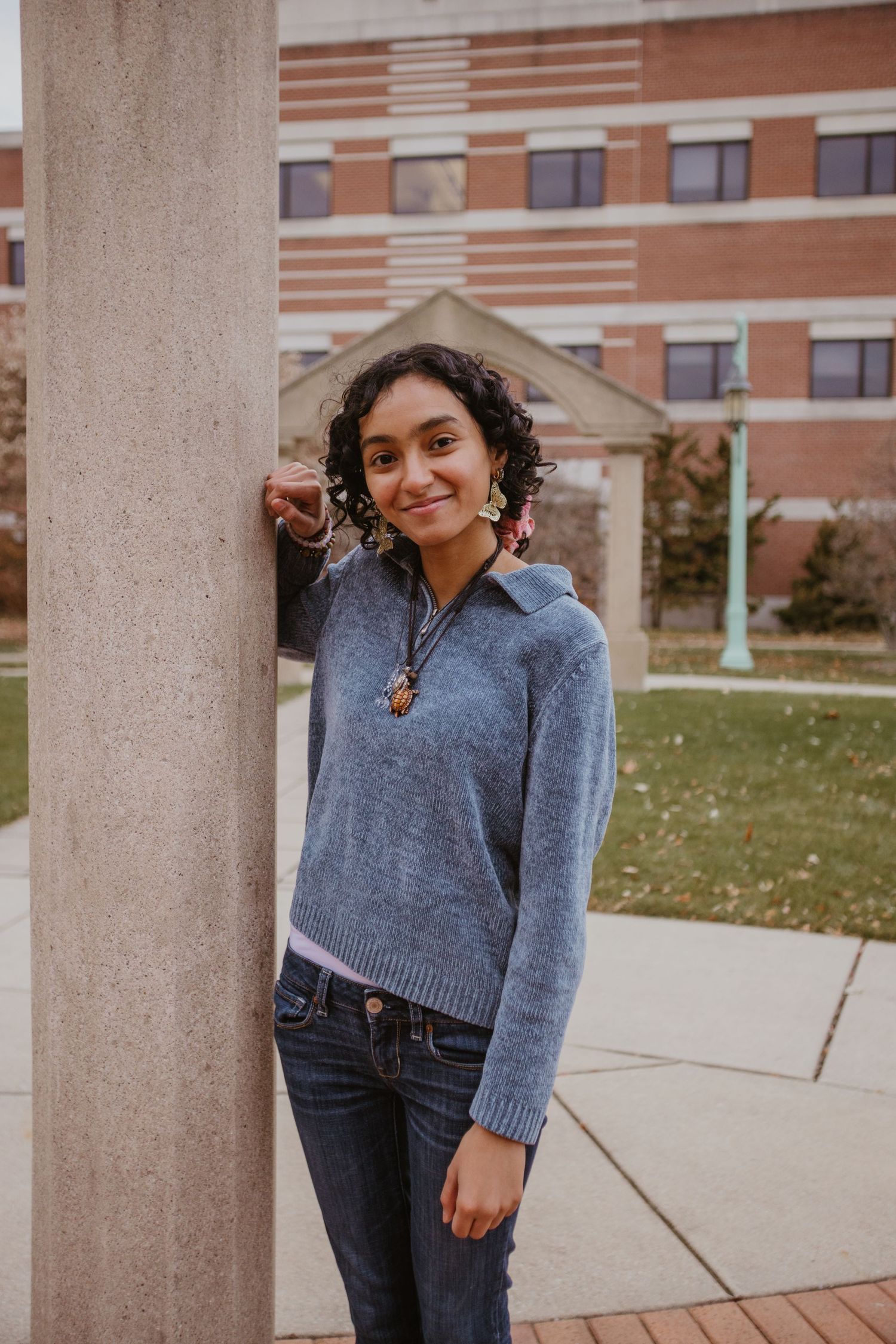 Someone in a cozy gray sweater and jeans poses casually near concrete columns on a college campus.