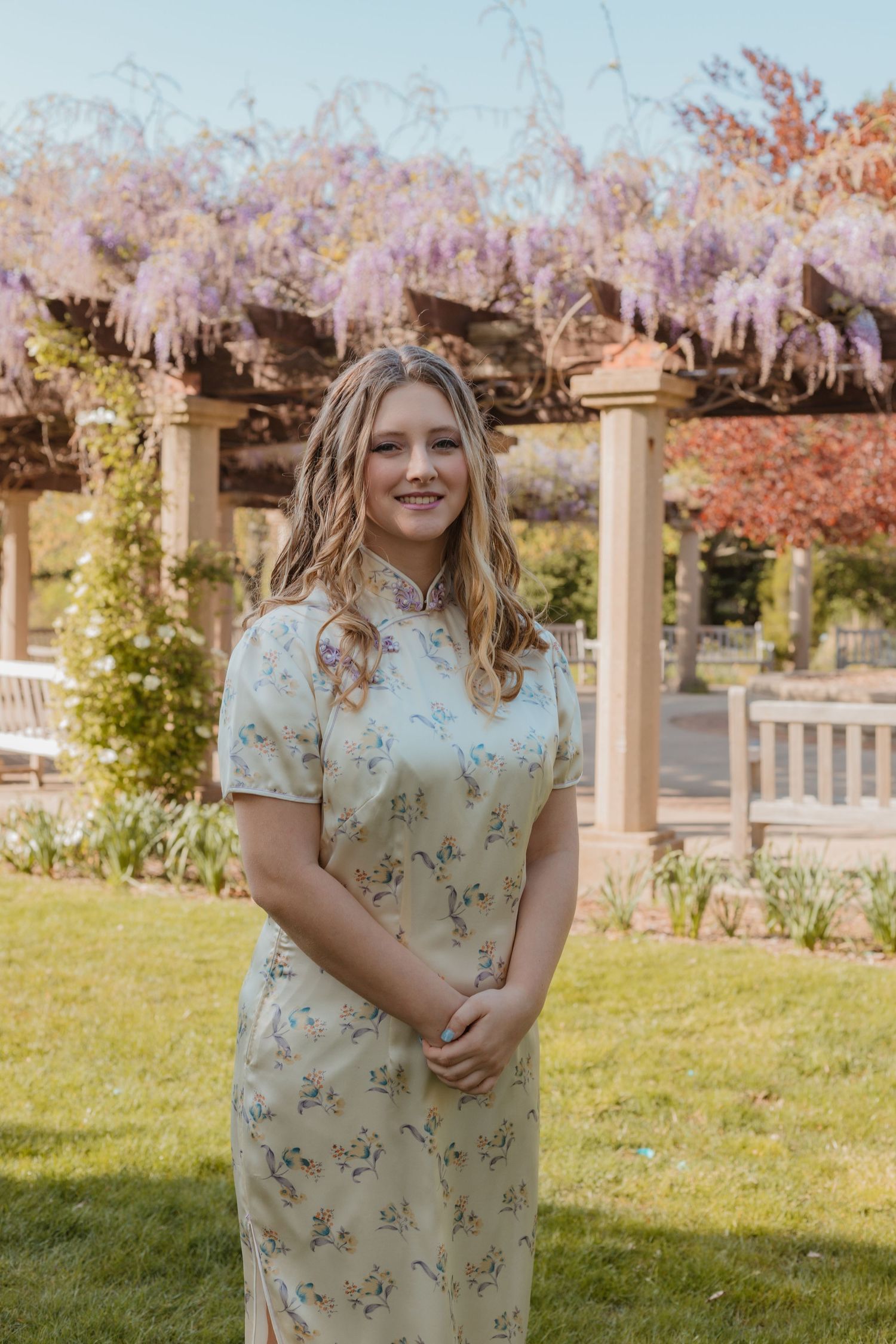 A person in a floral dress stands under blooming cherry blossom trees in a garden setting during spring.