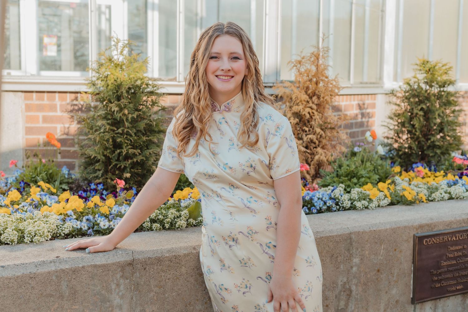 Someone in a floral dress poses outdoors near colorful flowers and a stone wall on a sunny day.