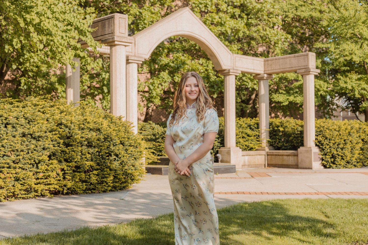 A person in a floral dress stands in a scenic garden pathway with white architectural columns and lush greenery.