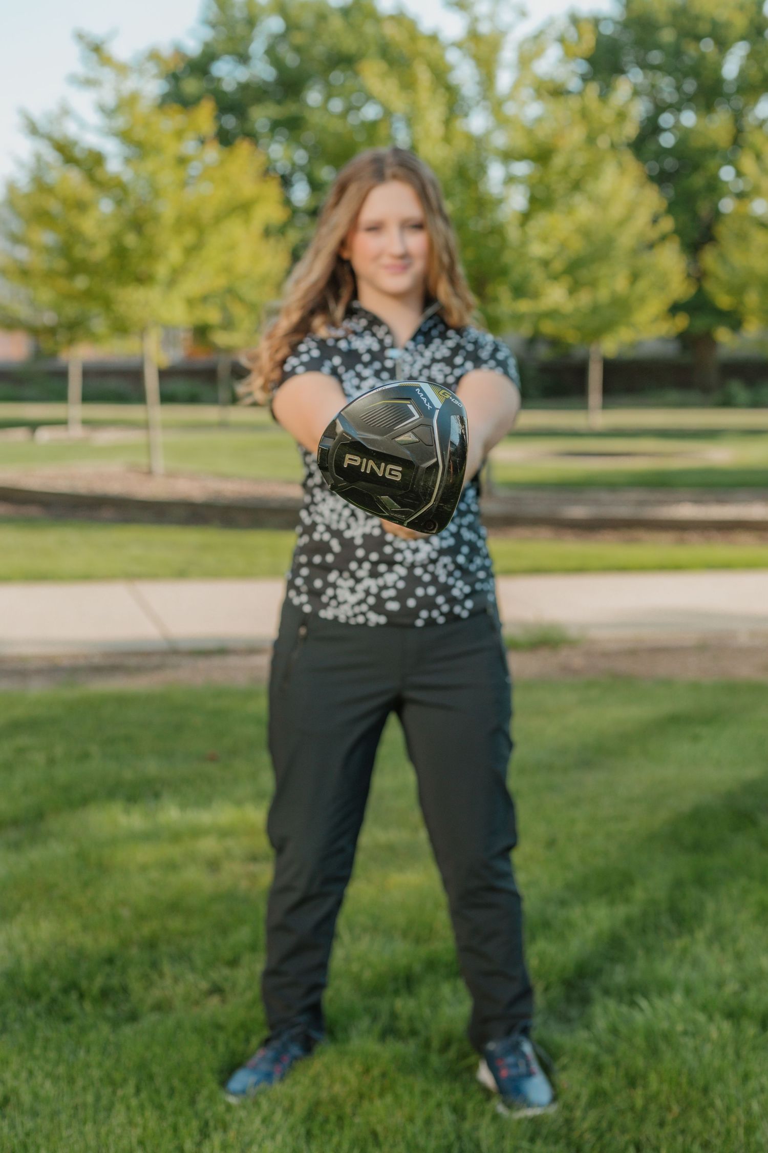 Person wearing patterned top and dark pants poses in grassy outdoor area with trees in background.