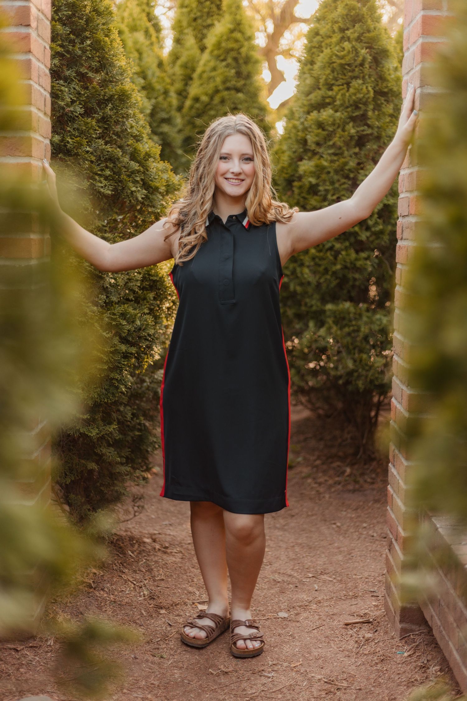 A person in a black sleeveless dress poses joyfully between evergreen trees in an outdoor garden setting.