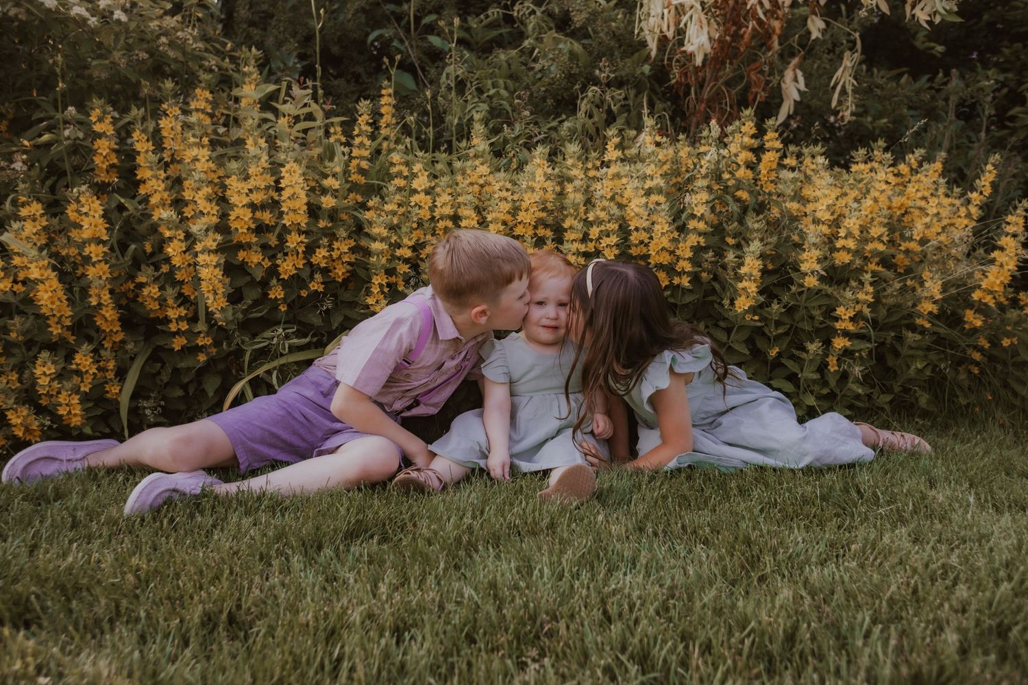 A couple in casual attire lays together on grass during golden hour surrounded by yellow wildflowers.