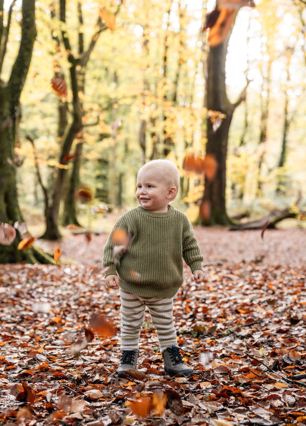 Small child in brown sweater and striped pants plays joyfully among falling autumn leaves in a forest setting.