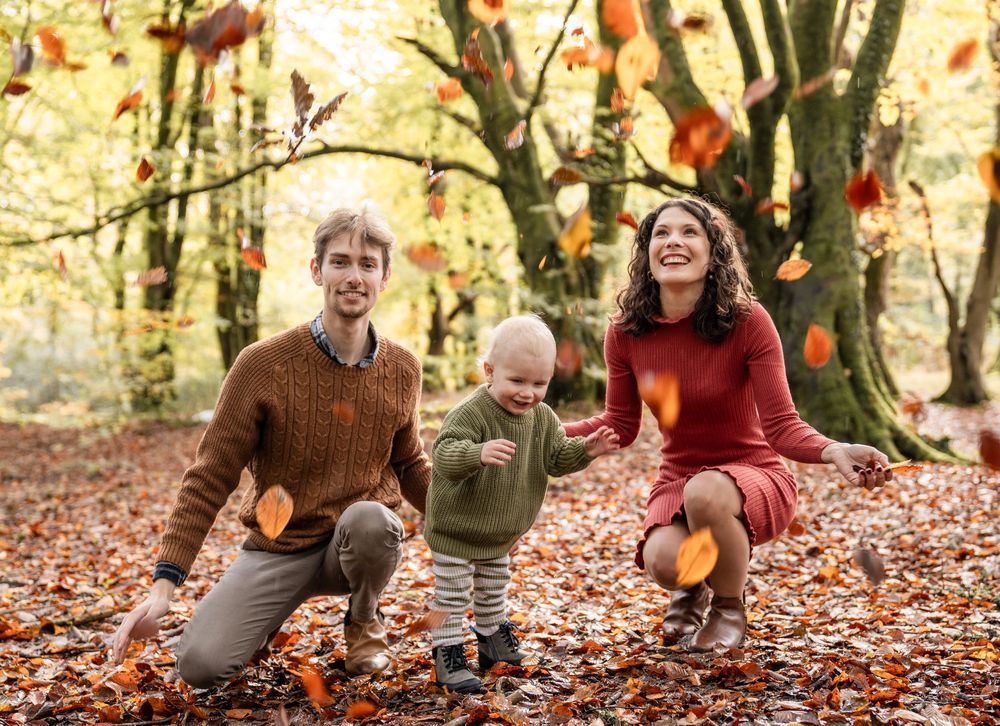 A family plays together in autumn forest with falling orange leaves around them in golden sunlight.