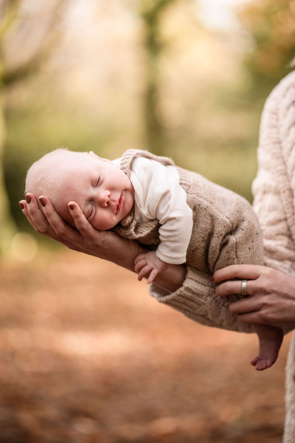 A gentle hand cradles a sleeping newborn baby wearing a white onesie and brown knit sweater in soft natural light.