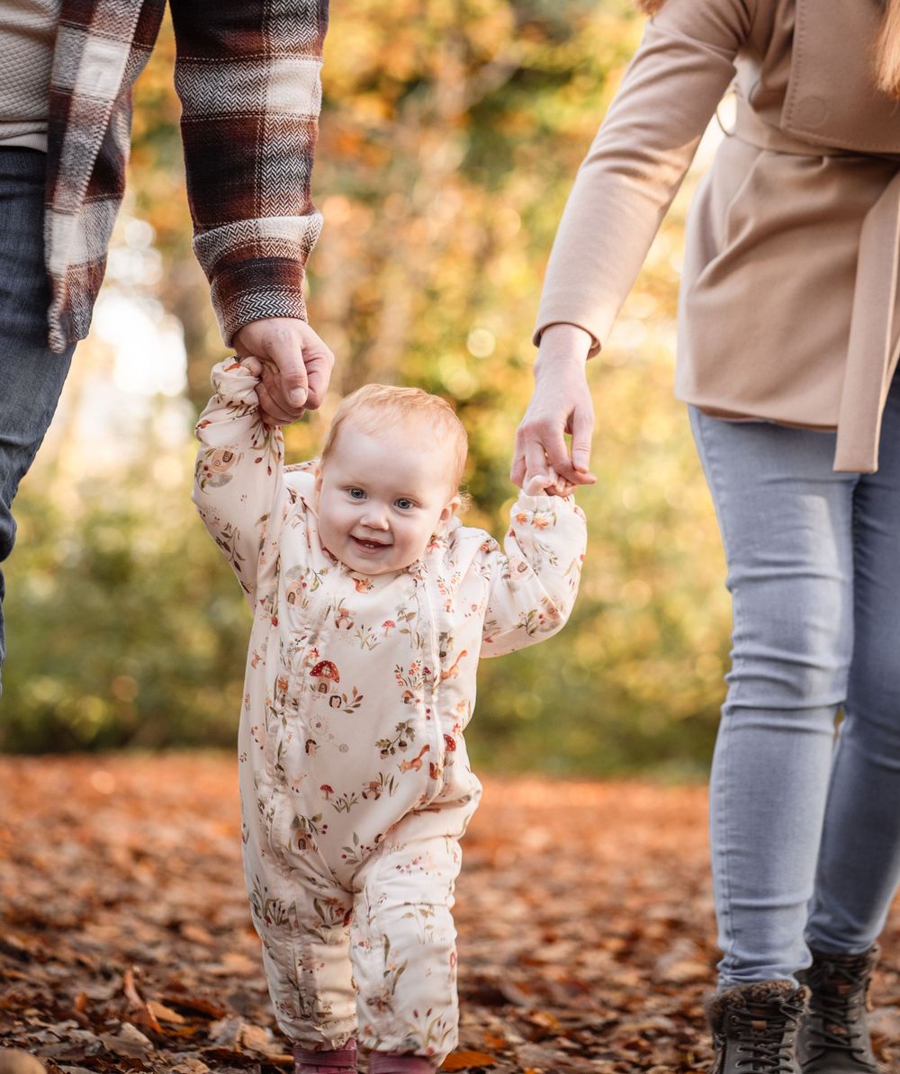 A child plays in fallen autumn leaves while wearing a floral outfit and being supported by adults in a park setting.