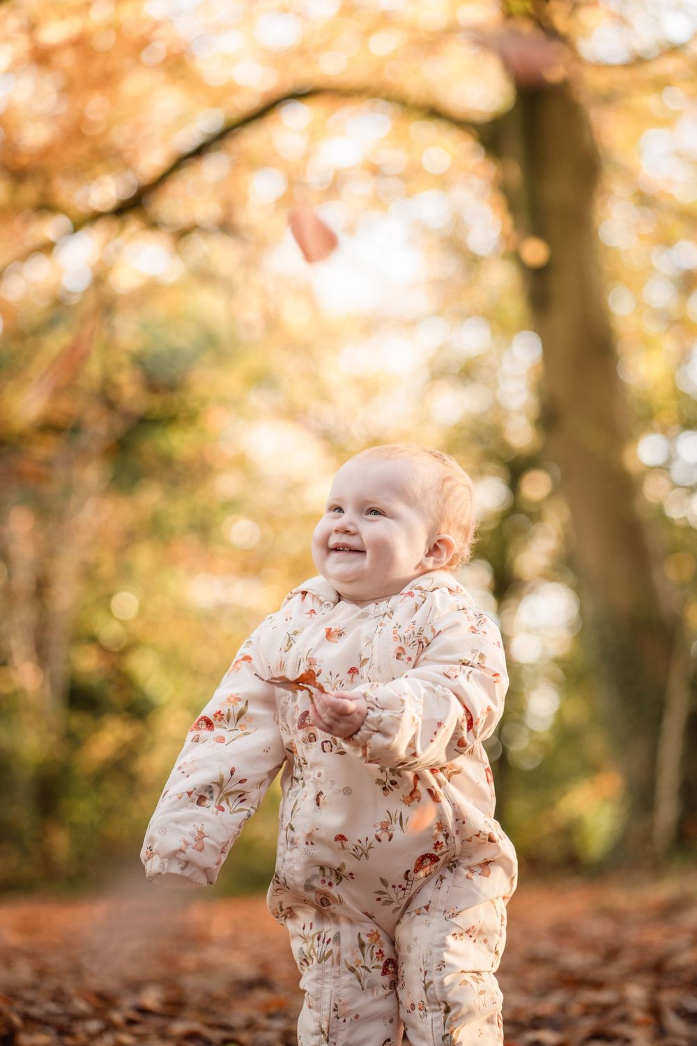 A small child in a floral outfit plays joyfully among fallen autumn leaves in a sunlit forest setting.