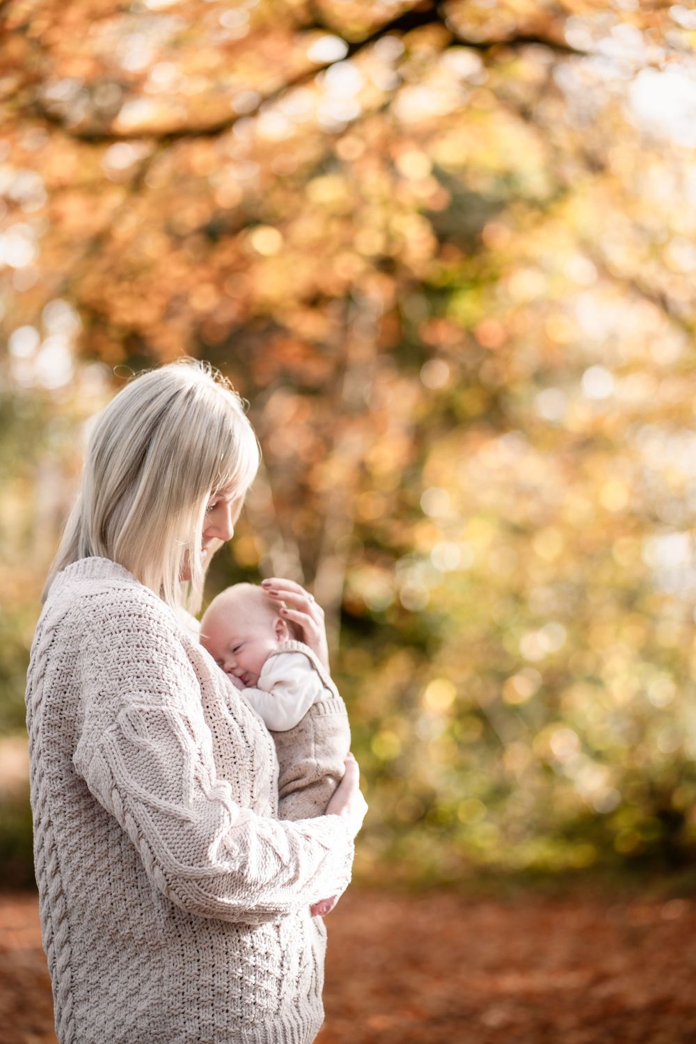 A parent in a cozy beige sweater holds and gazes lovingly at their baby against an autumn backdrop with golden bokeh.