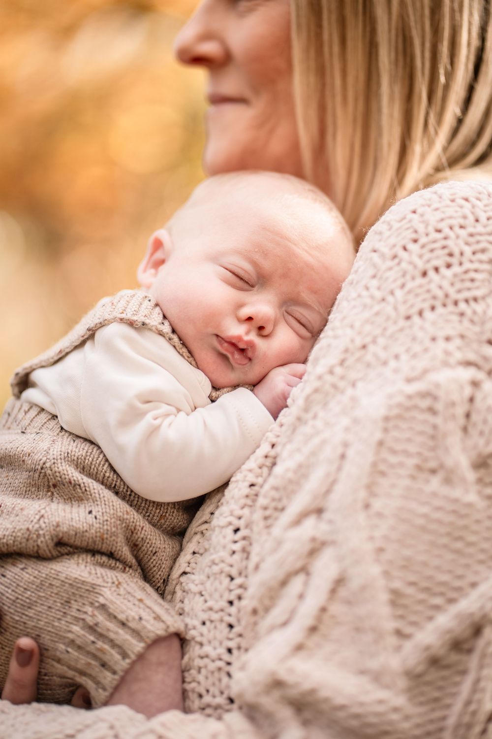 A sleeping newborn baby swaddled in white rests peacefully against a knit sweater in warm autumn lighting.