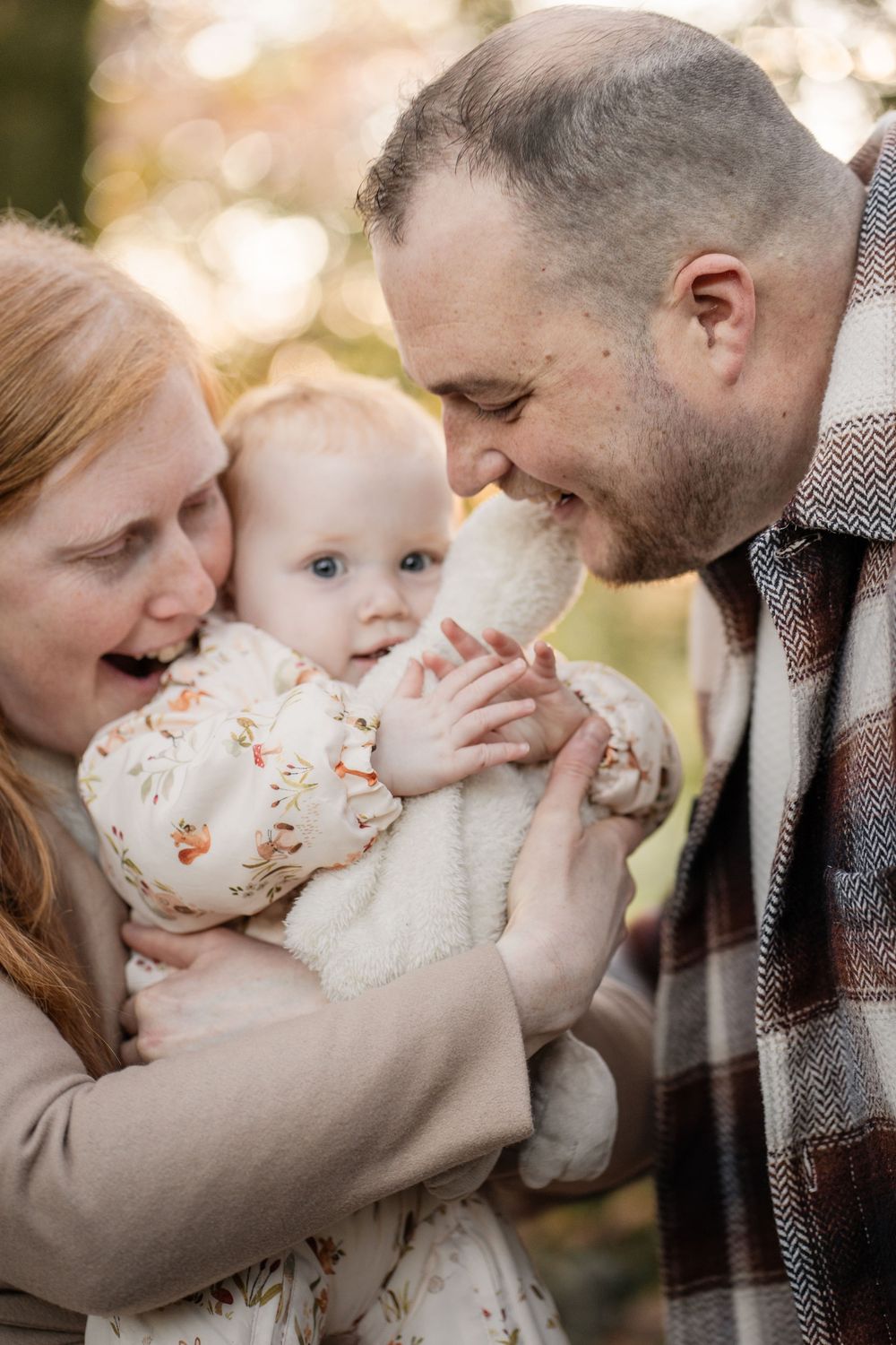 A tender family moment captured outdoors with a baby in a floral outfit being lovingly embraced by parents.