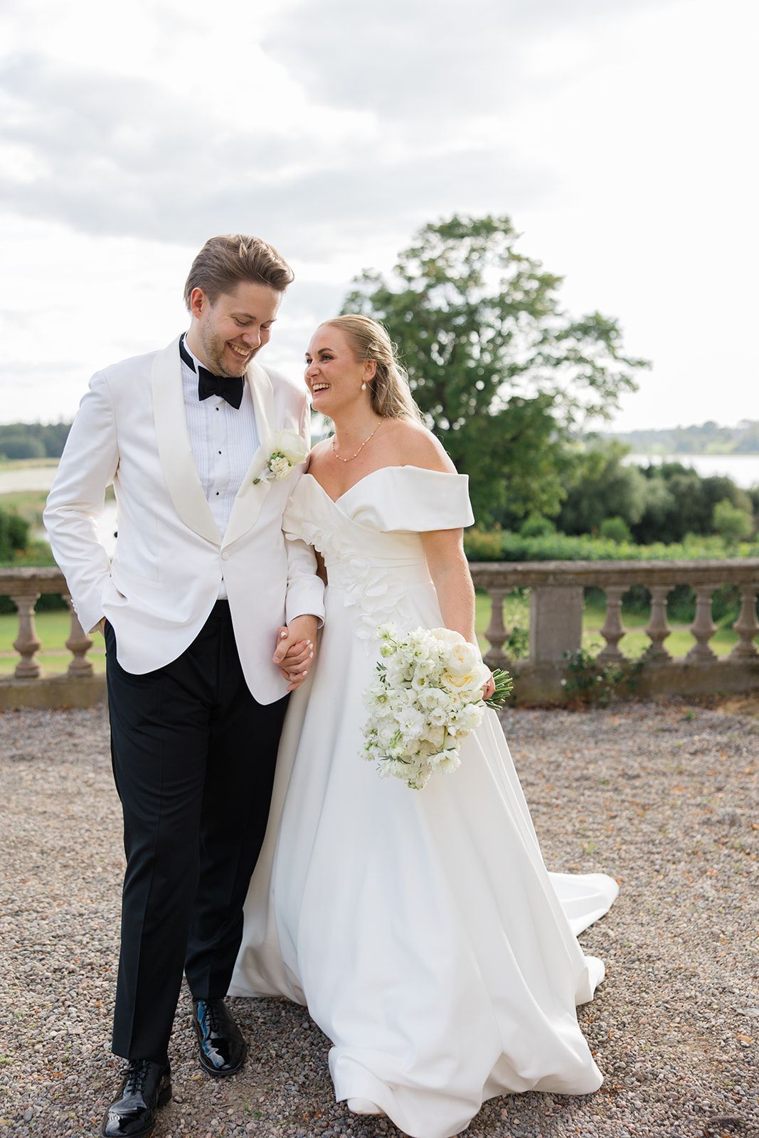 A couple in wedding attire stands on a gravel path in front of a grand yellow mansion with manicured hedges.