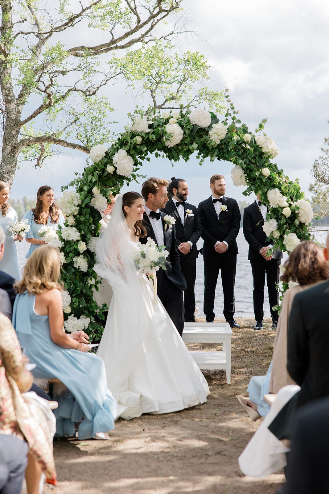 An outdoor wedding ceremony under a lush floral arch with greenery and white roses overlooking the water.