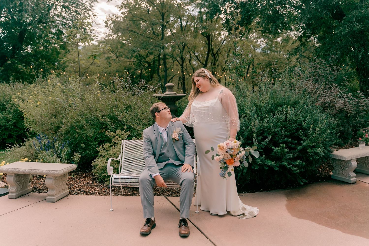 A couple sits on a rustic wooden bench in a lush garden setting during a wedding ceremony.