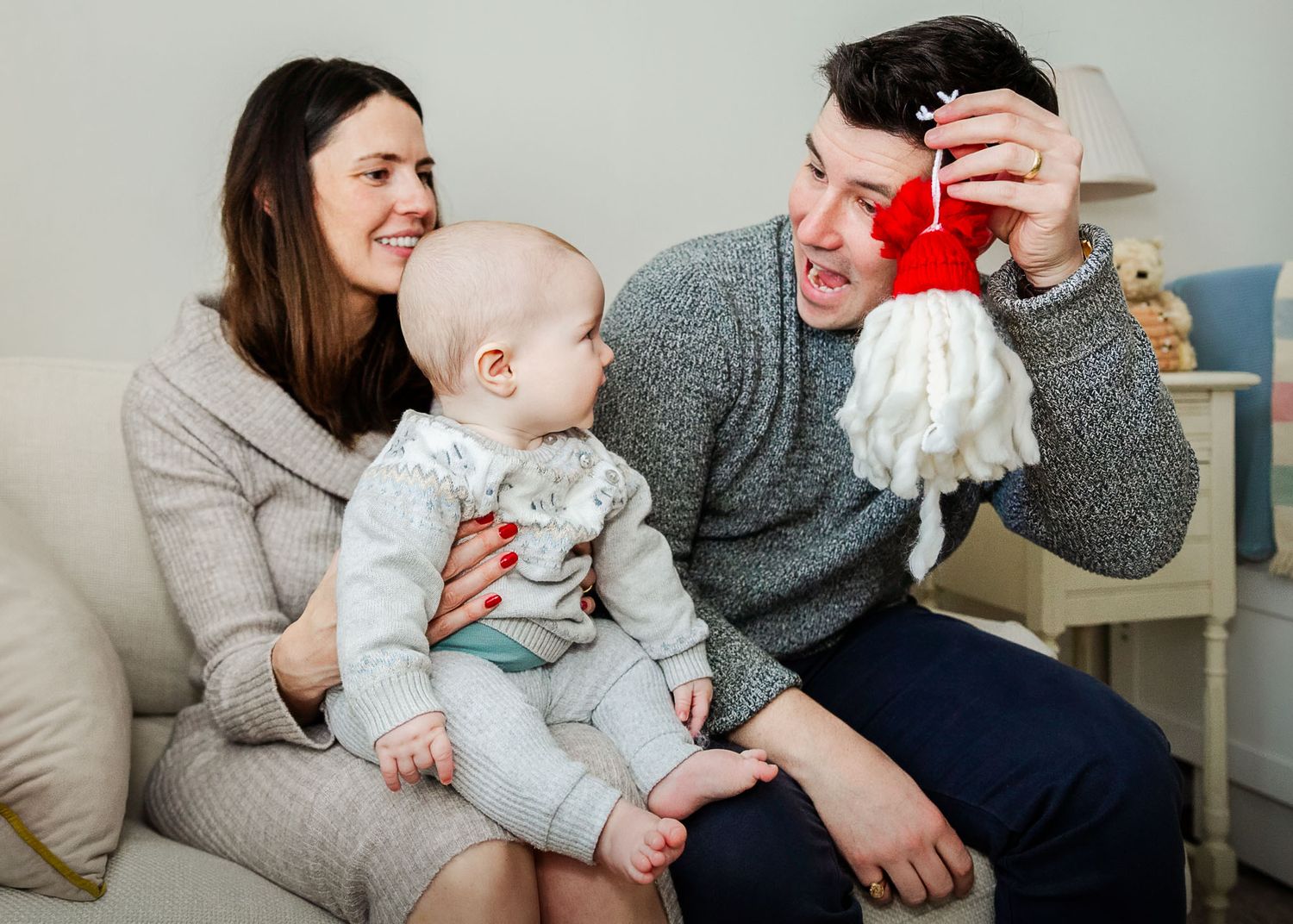 A baby sits with his mum and looks at his dad who is holding a gonk up and smiling. The family look relaxed. Christmas photoshoot in Bristol