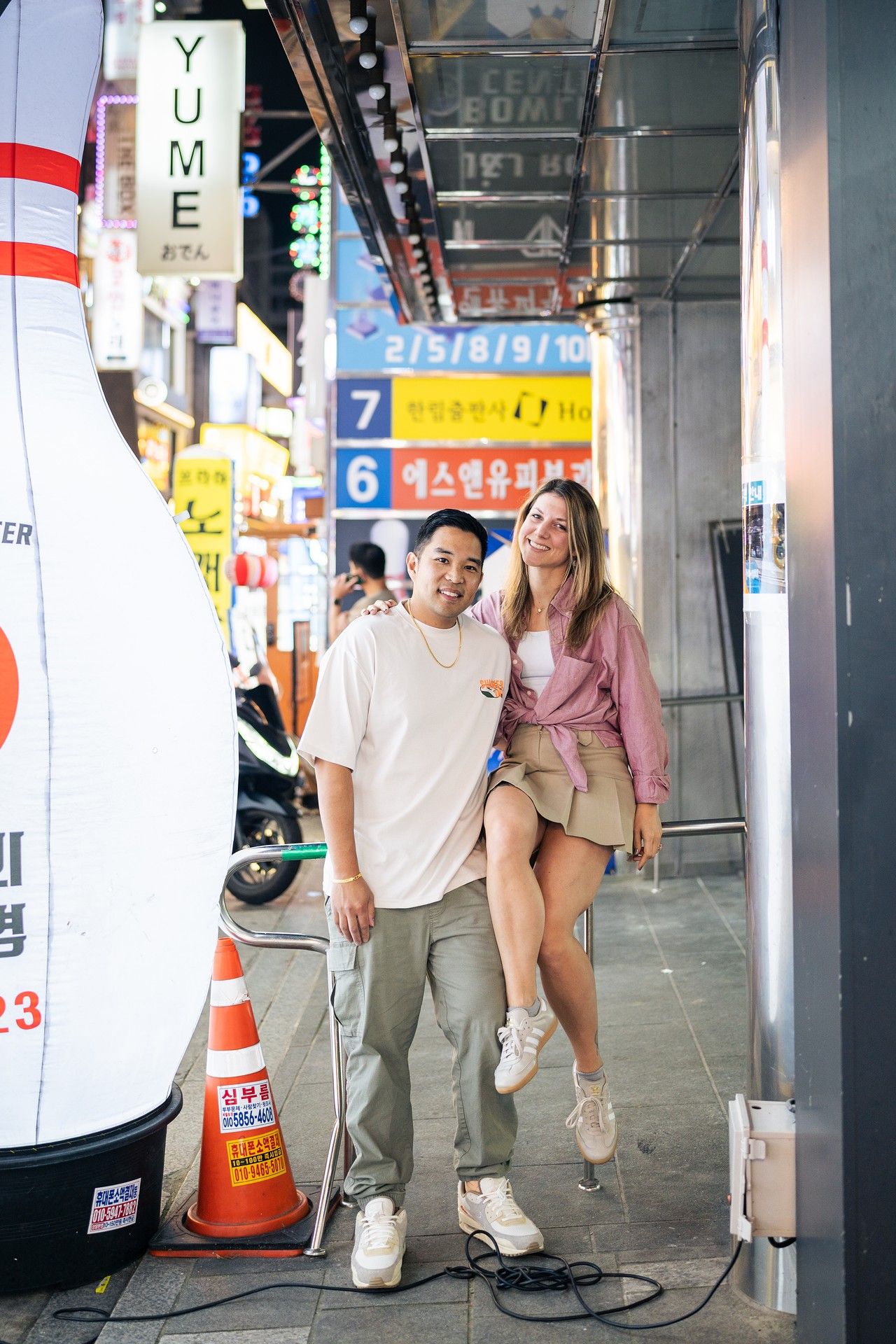 Couple walking together on a city street with neon signs and urban architecture in the background.