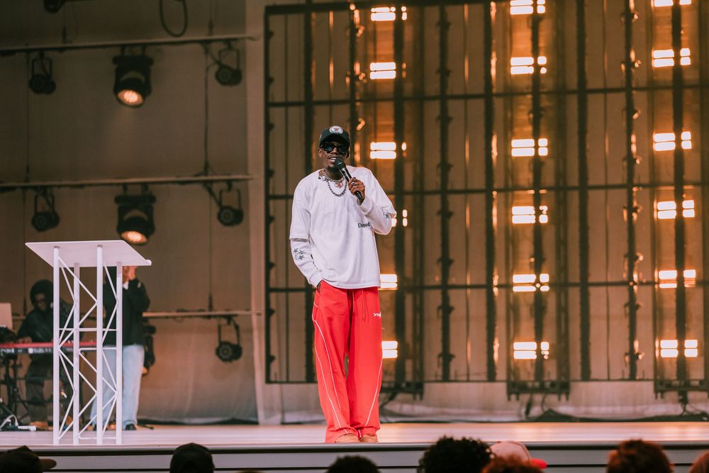 Performer in white hoodie and red pants speaking into a microphone on a church stage.