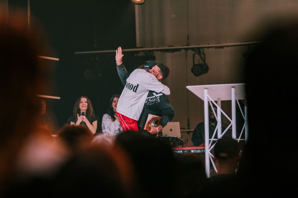 Two people hugging on a dimly lit stage during a live event with an audience.