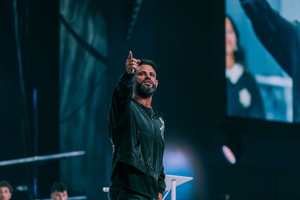 Bearded male speaker points upward on a large stage with screens during a live conference event.
