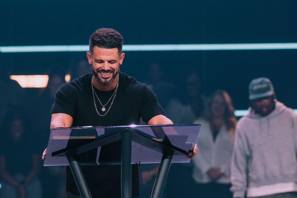 Smiling man in black shirt laughing at a clear podium on a church stage.