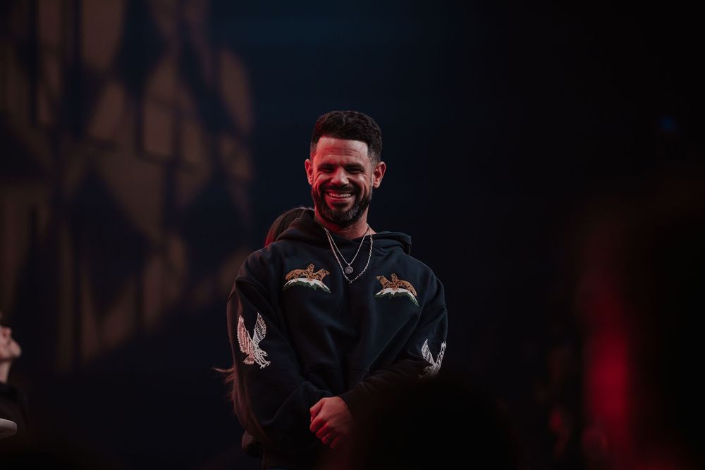 Smiling man in black embroidered hoodie stands on stage under warm spotlight at a live event.