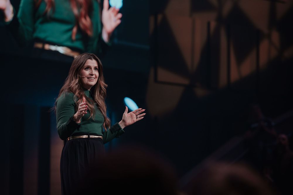 Woman in green outfit smiles and claps while speaking on a dramatically lit stage during a live event.