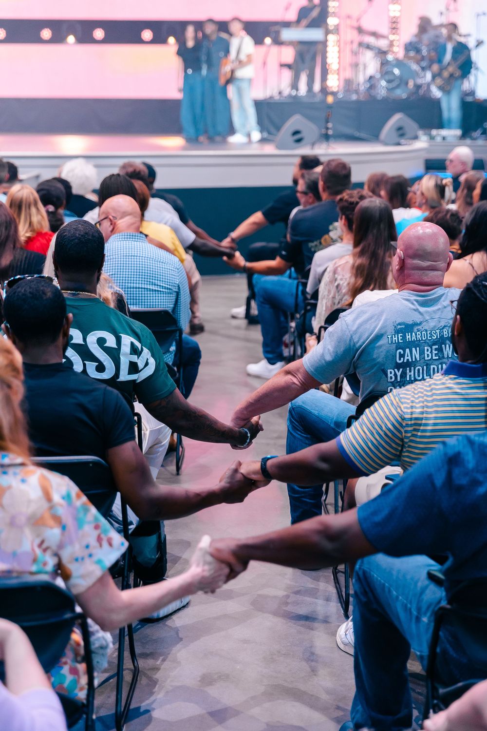 Diverse congregation holds hands in prayer at a live worship service with a band performing on stage.