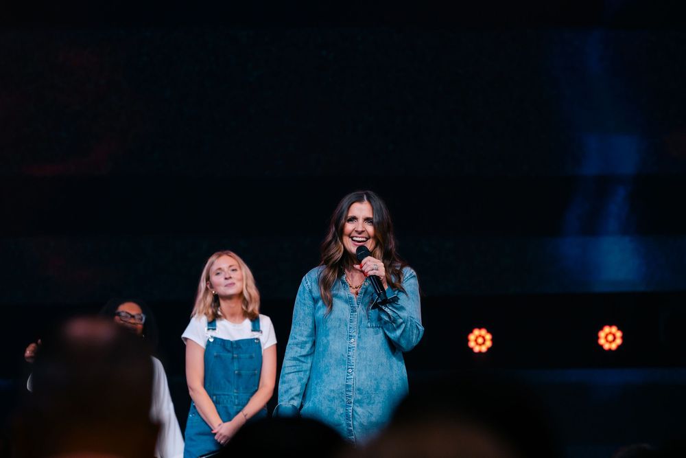 Woman in denim jacket speaks into microphone on stage with young girl in overalls beside her.
