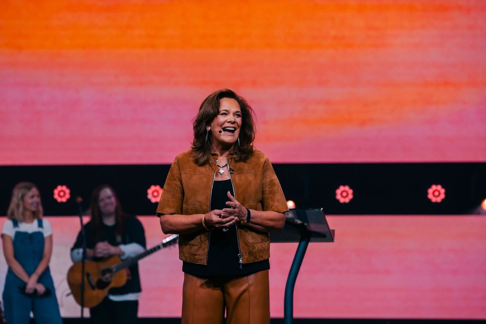 Woman in brown jacket speaks at podium on stage with orange backdrop and guitarist behind her.