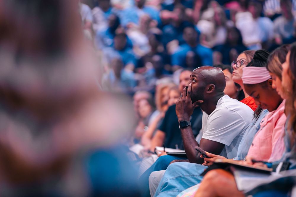 Diverse audience members seated in a crowded arena, attentively watching an event, with a man in a white shirt in focus.