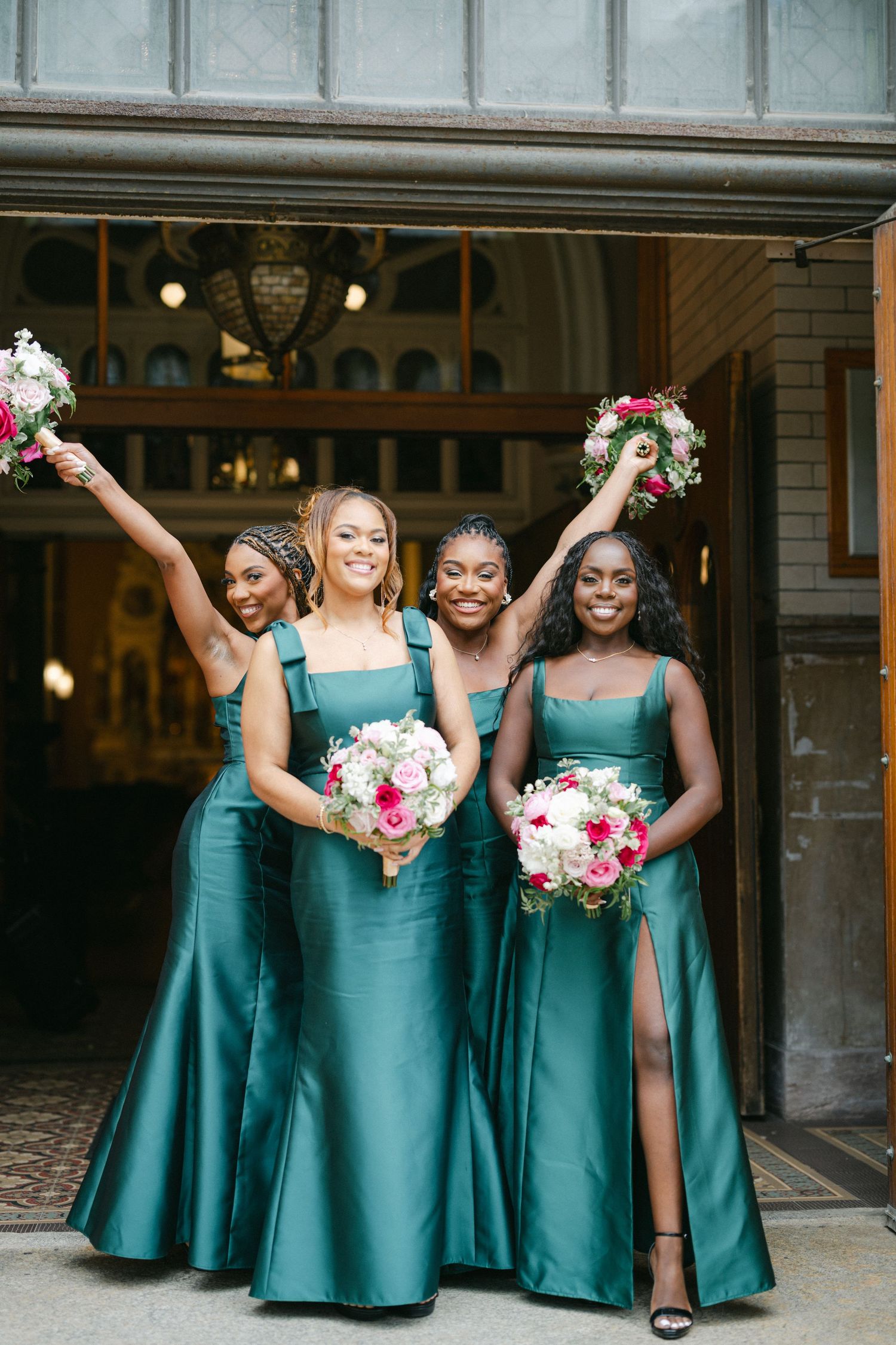 Bridesmaids in matching teal satin gowns hold pink and white bouquets while posing together at a wedding venue.