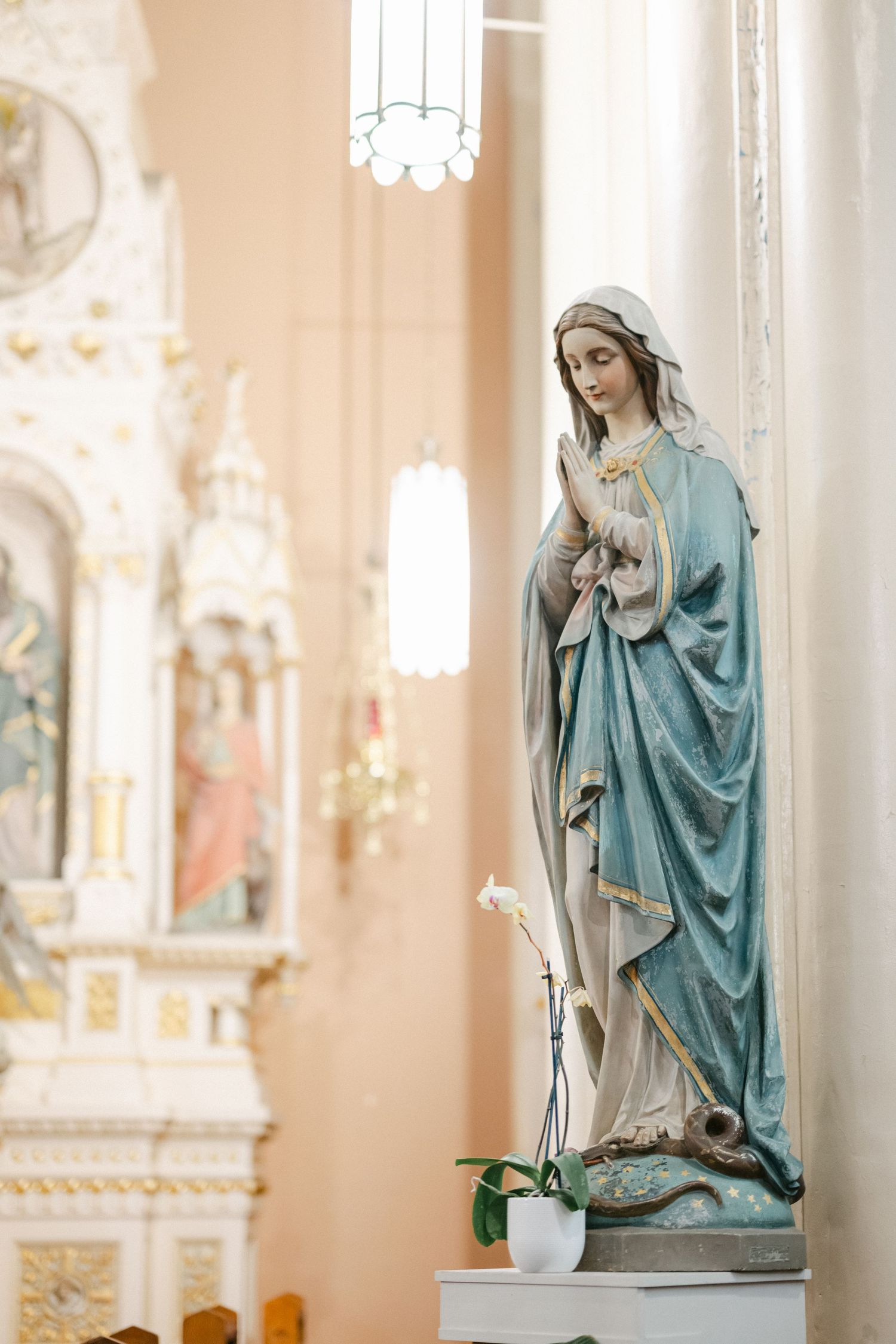 A religious statue of Mary in blue robes stands inside an ornate church with soft lighting and decorative details.