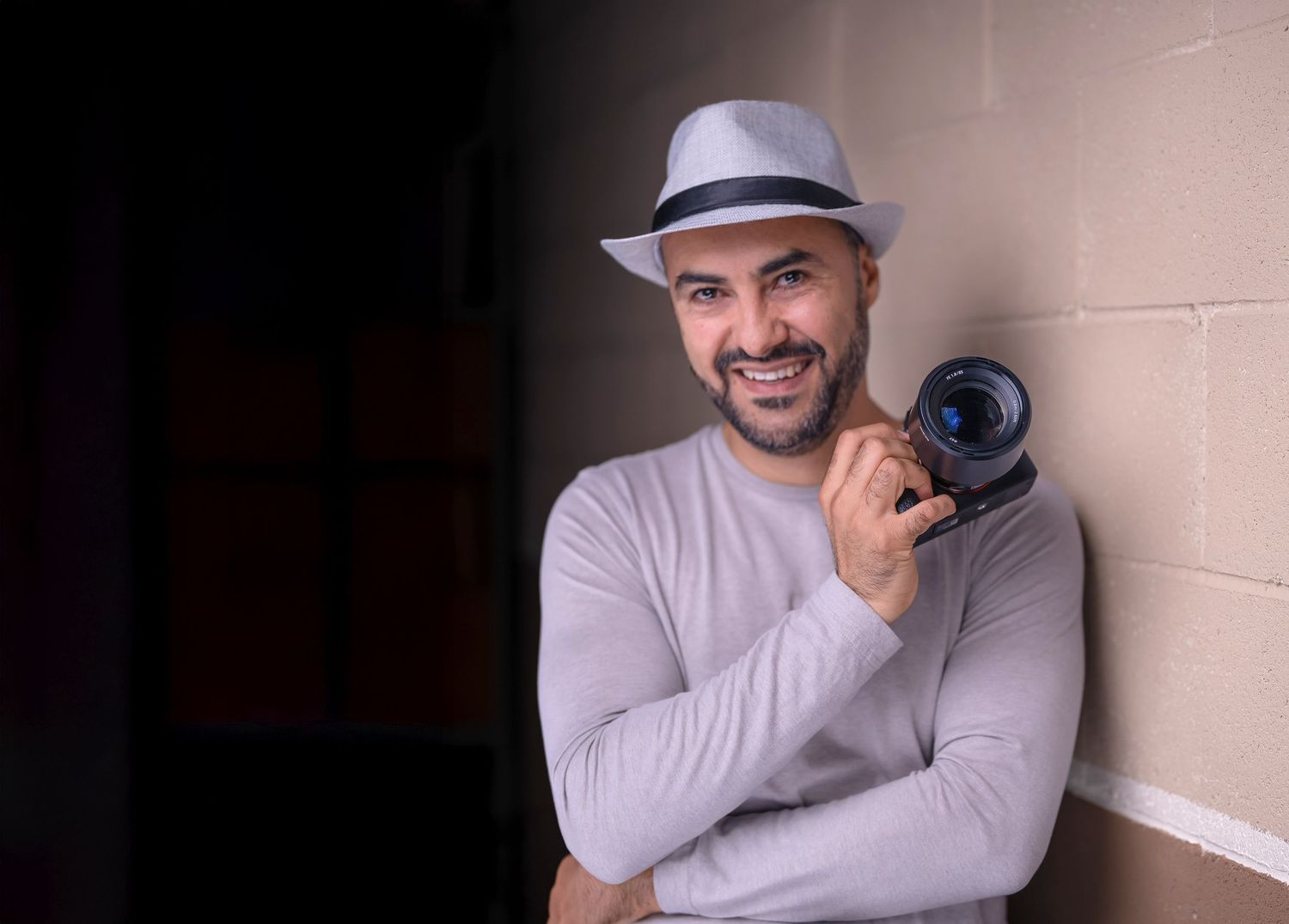 A photographer in a black shirt and white hat smiles while holding a camera at an outdoor event.