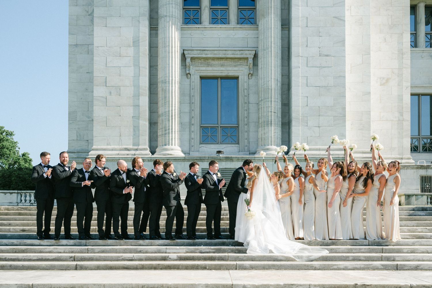 Wedding party poses on grand white marble steps of historic courthouse building in elegant formal attire.