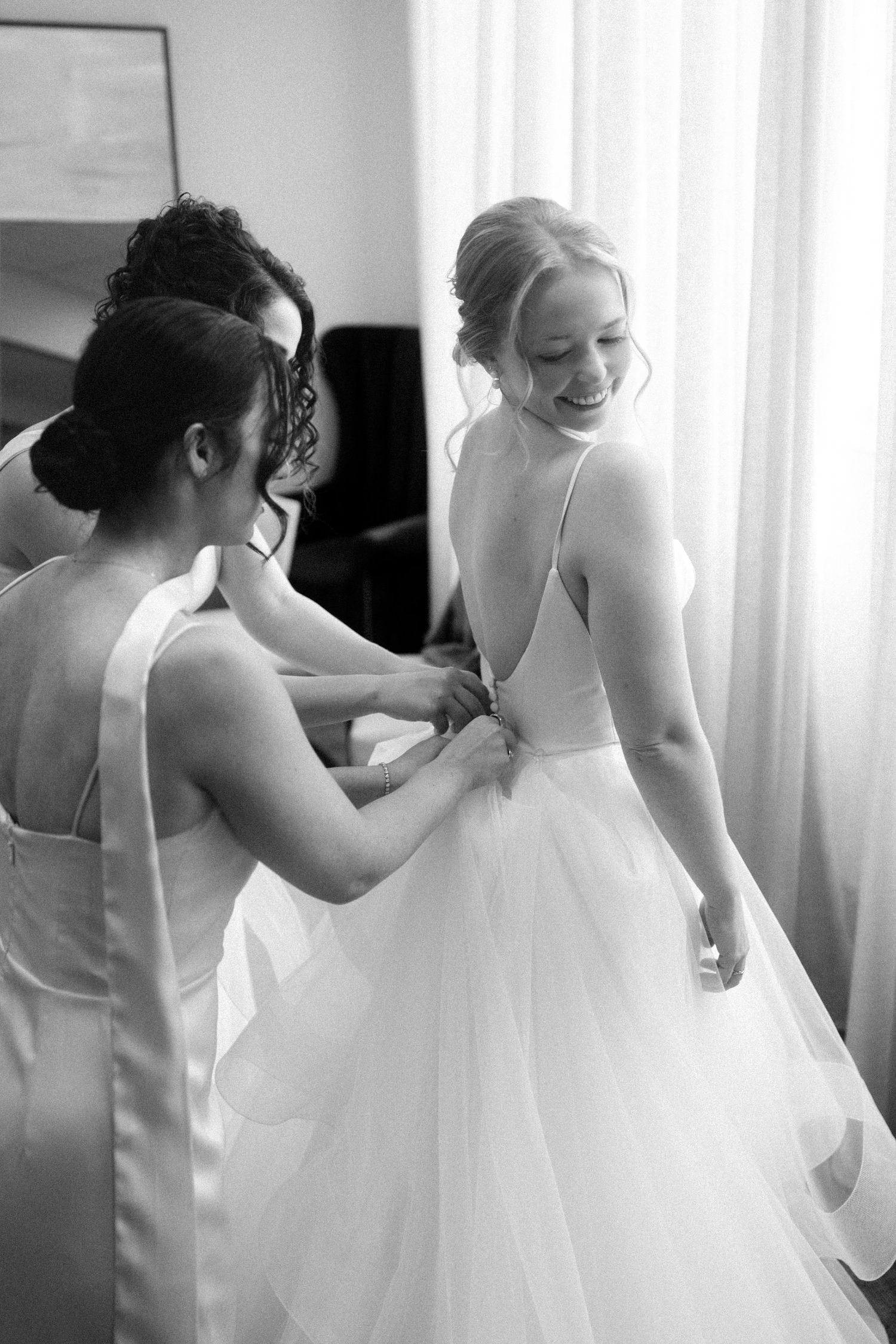 Bridesmaids help adjust the bride's white wedding gown in a sunlit room.