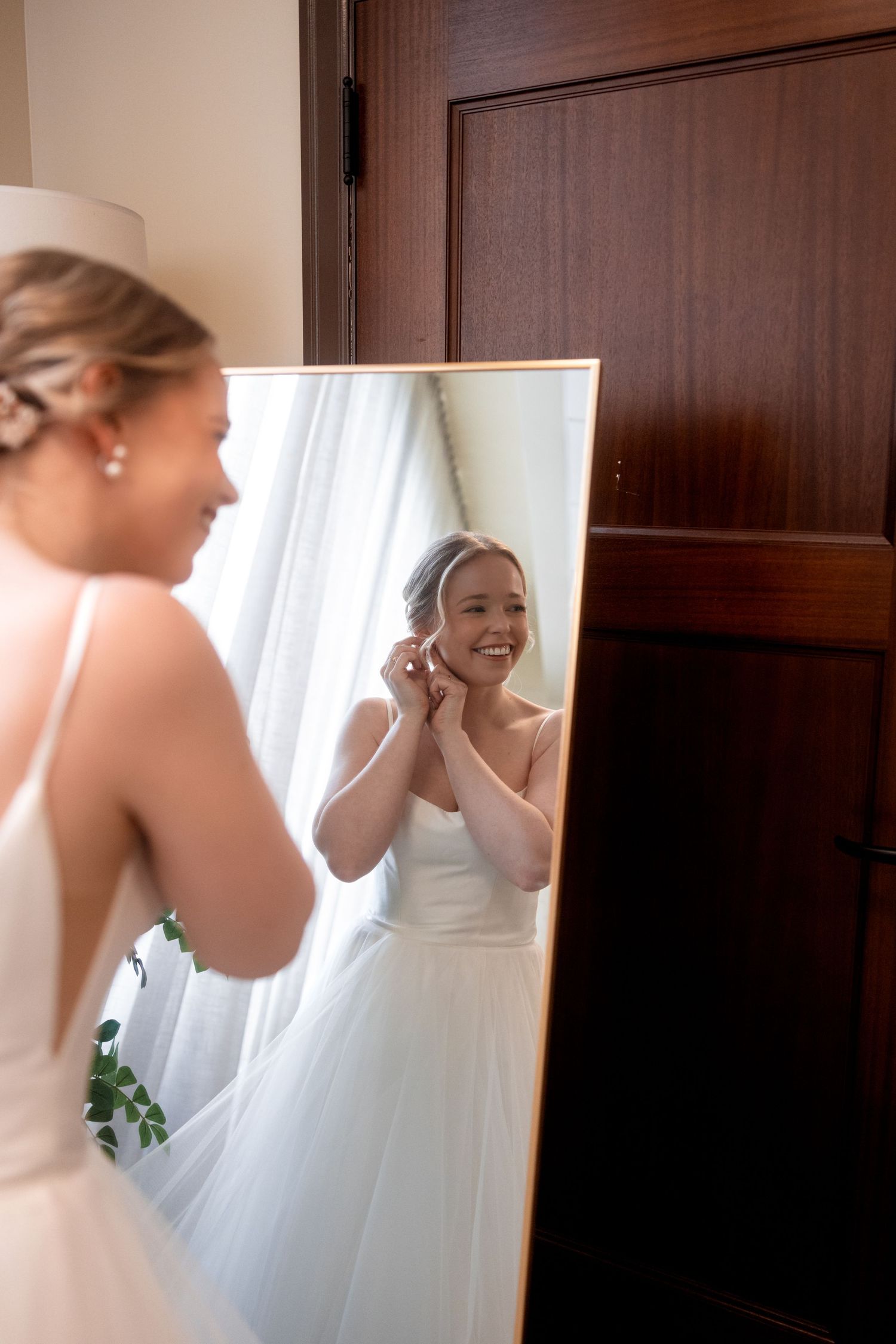 A bride in a white wedding dress puts on earrings while looking in a mirror during wedding preparations.