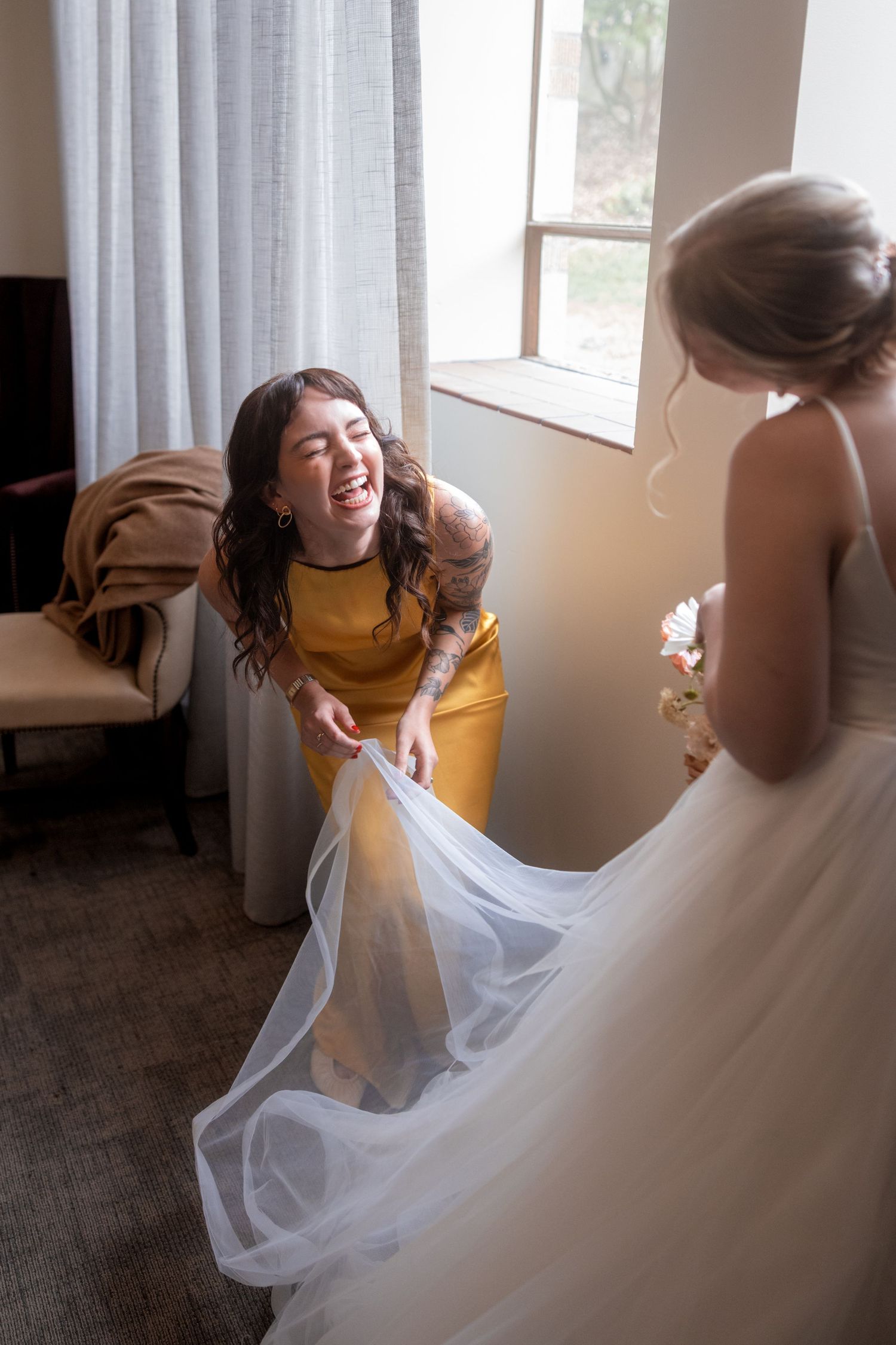 A joyful moment shared between a bride in white and friend in yellow as they laugh while handling the wedding dress.