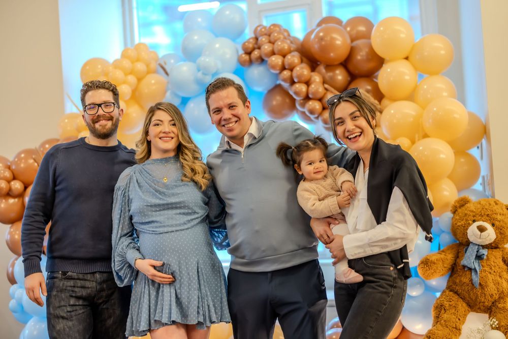 A group poses together at a celebration with blue and gold balloon decorations and a teddy bear in the background.