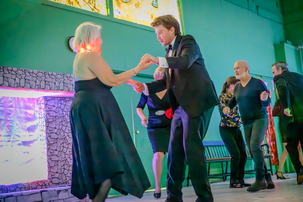 Couples dancing together during a social event with teal walls and decorative lighting in the background.