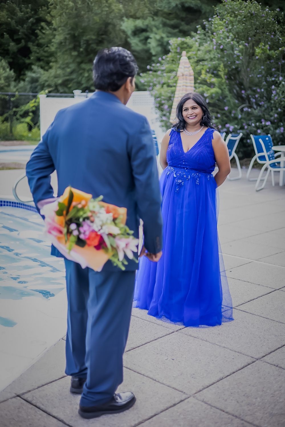 A person in a navy suit holds colorful flowers behind their back while facing someone wearing a flowing royal blue gown outdoors.