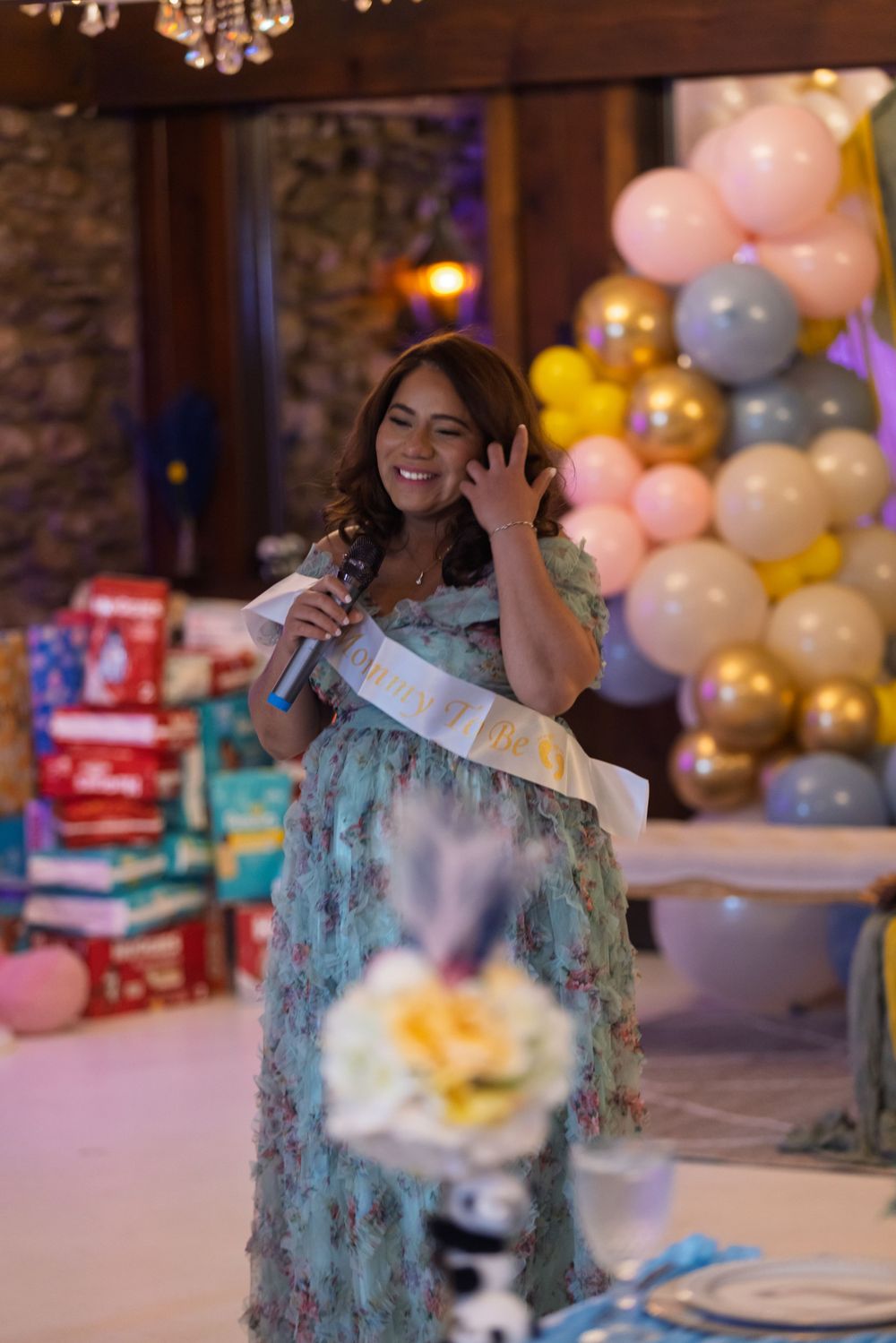 A baby shower celebration with pink and gold balloon decorations, wrapped gifts, and someone in a blue dress holding a microphone.