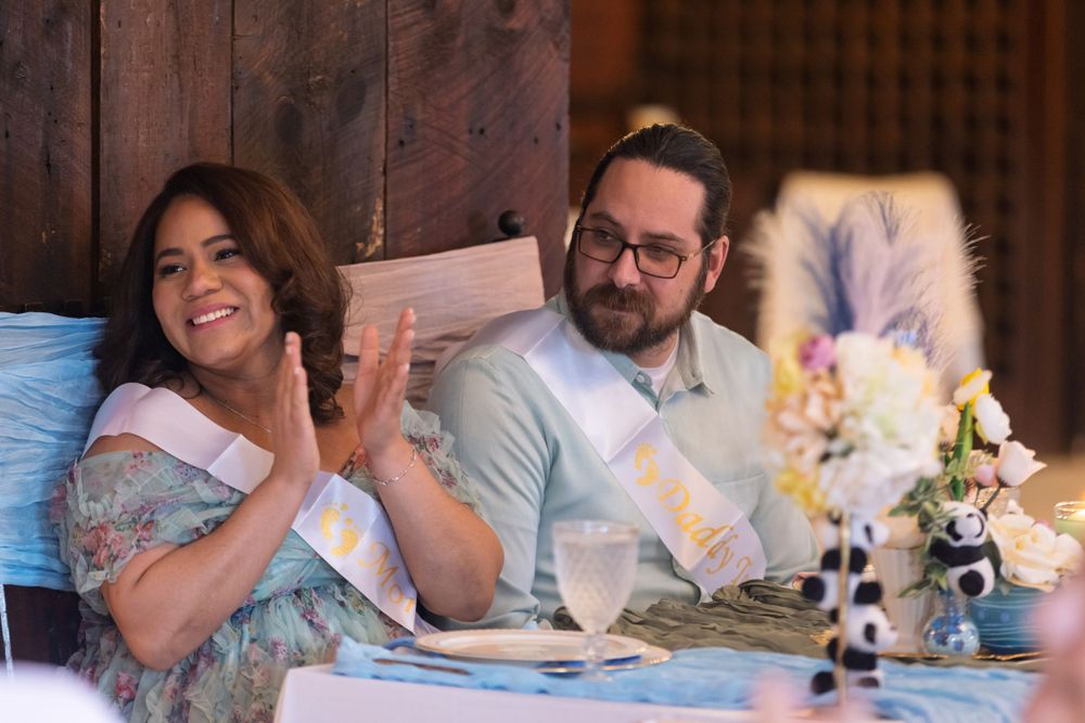 A couple smiling while seated together on a couch surrounded by books and flowers at an indoor celebration.