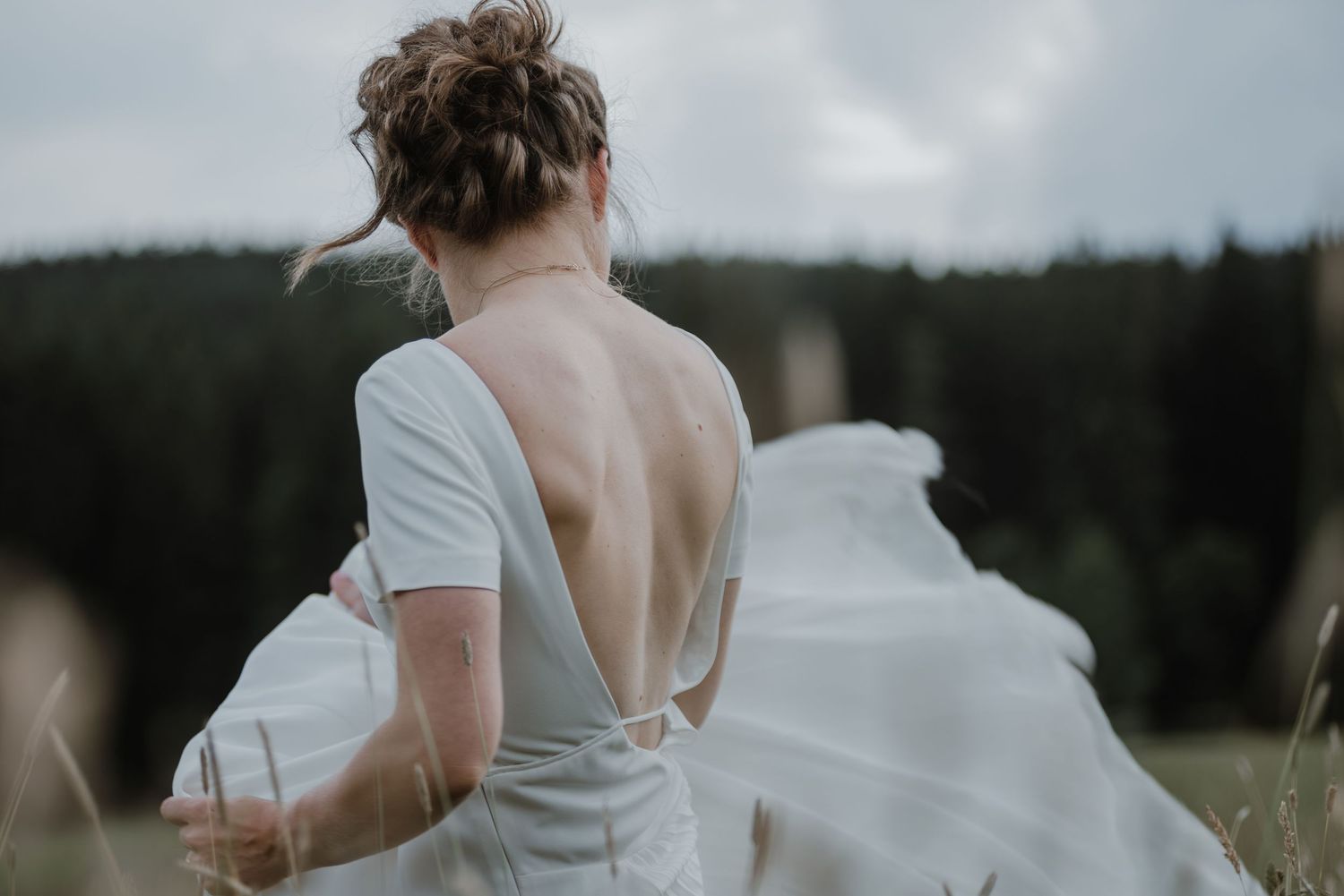 A flowing white dress with an open back design billows in the wind against a moody forest backdrop.