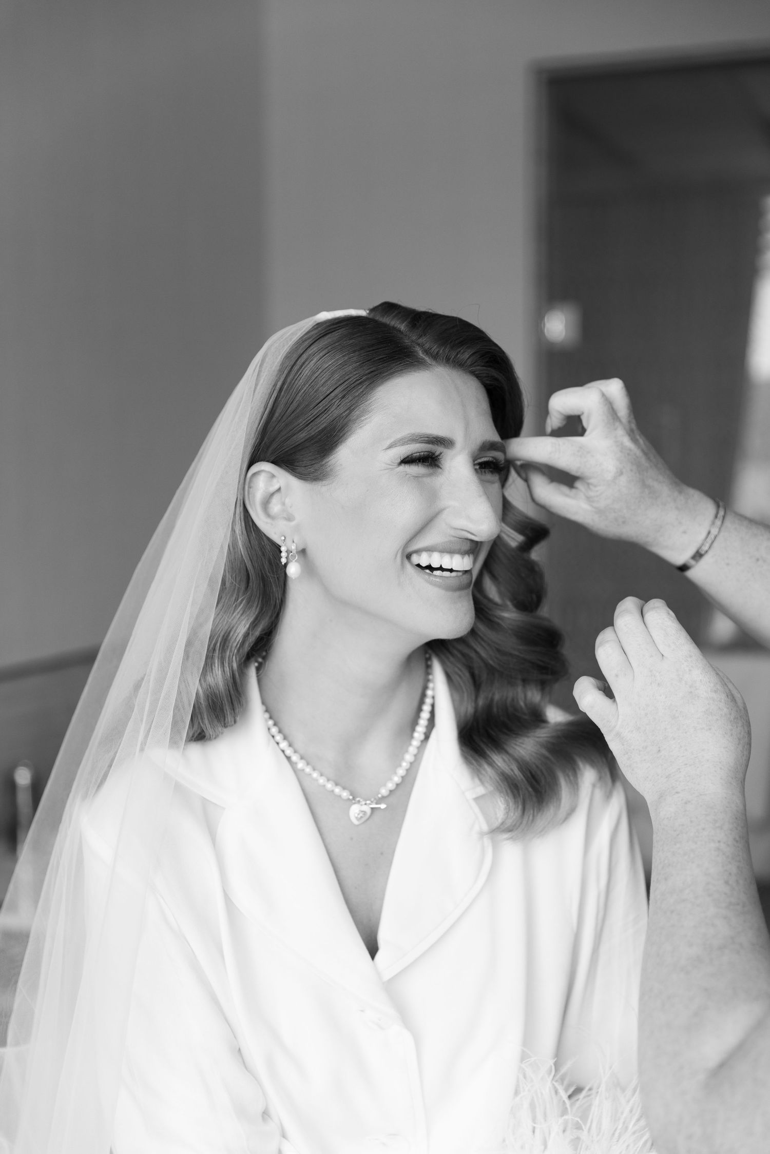 Black and white photo of bride in white robe getting makeup done while laughing during wedding preparations.