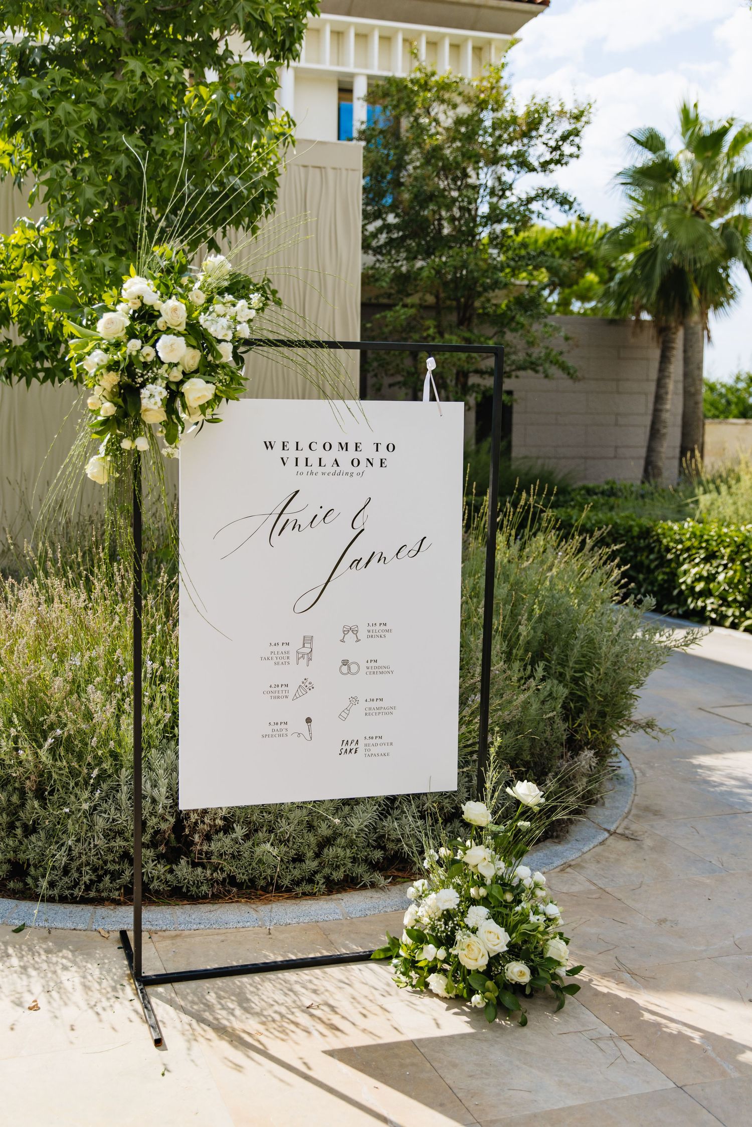 An elegant white wedding welcome sign with floral arrangements and palm trees in a garden setting.