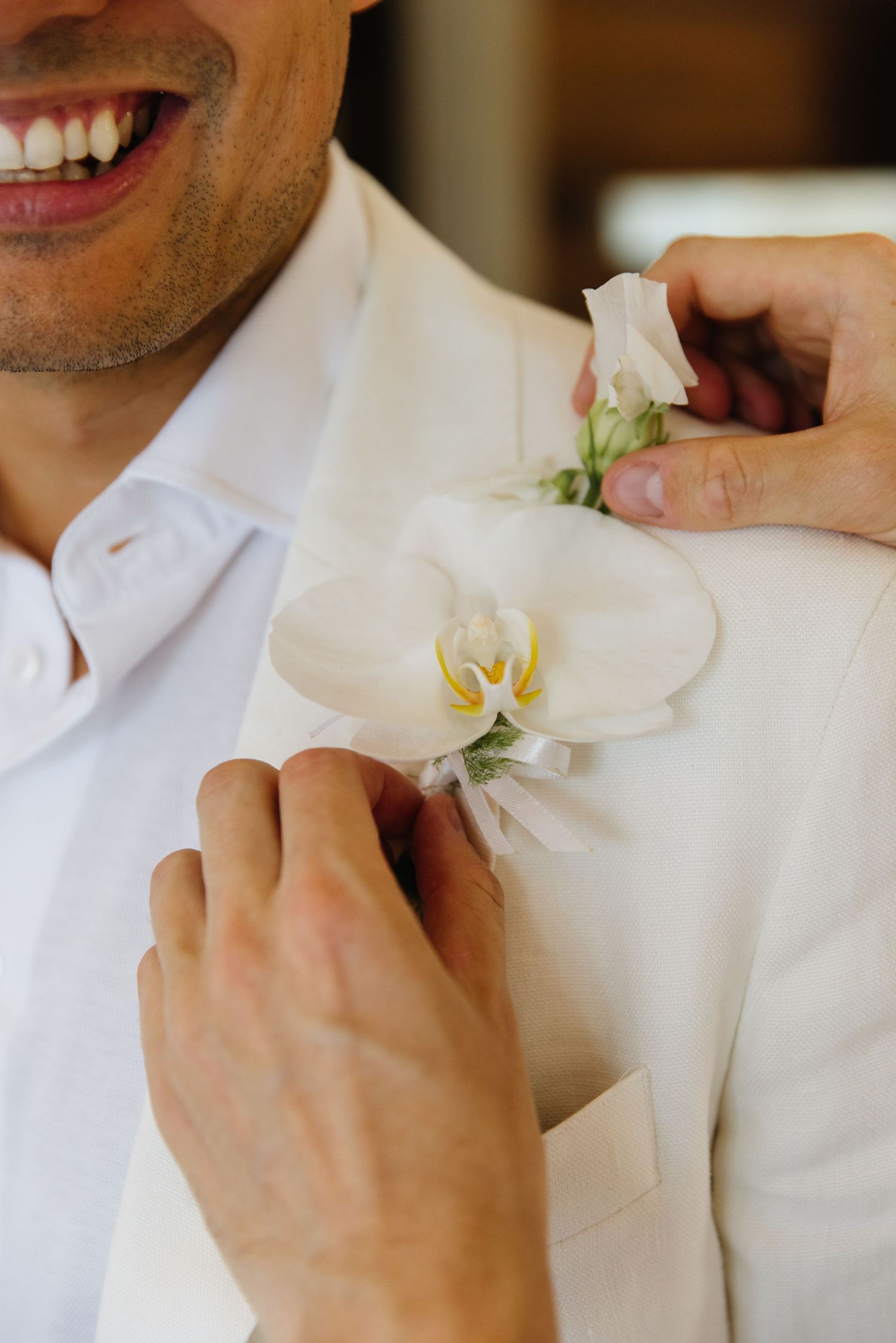 Hands pinning a delicate white orchid boutonniere onto a cream colored suit lapel at a formal event.