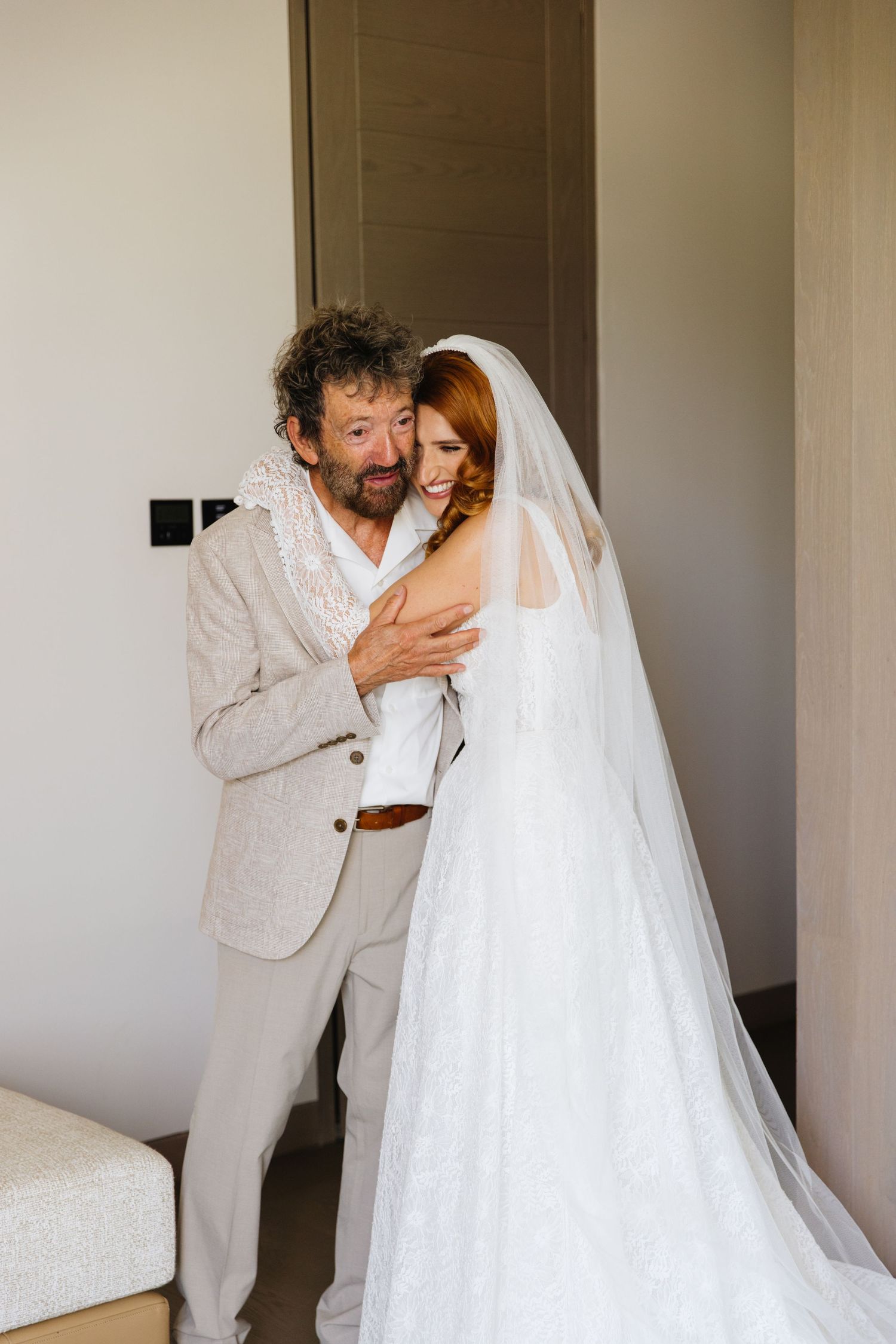 A tender moment captured at a wedding featuring a bride in a white lace gown with veil and companion in a light suit.