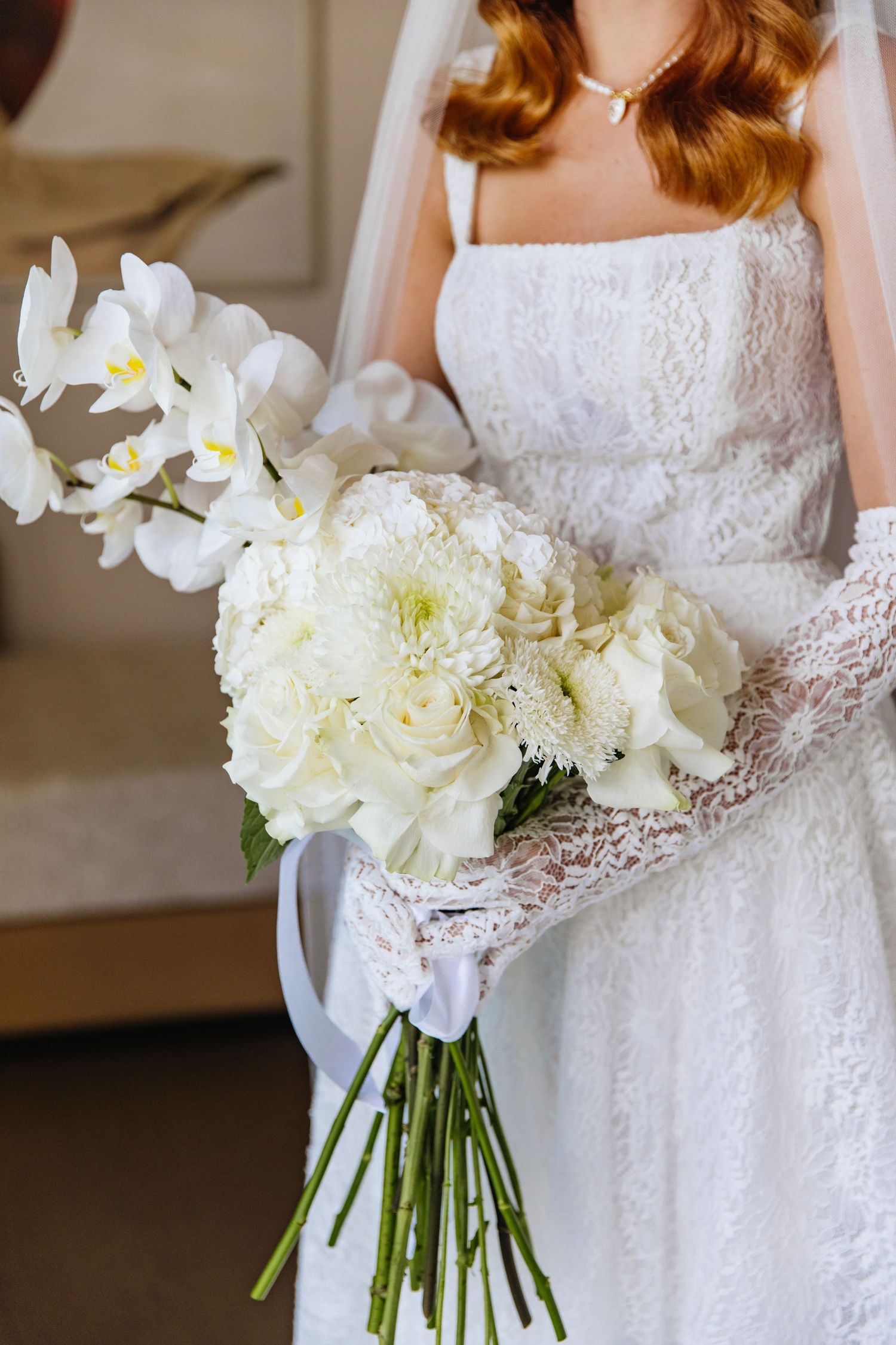 A bride in a white lace dress holds an elegant bouquet of white orchids and hydrangeas with delicate stems.