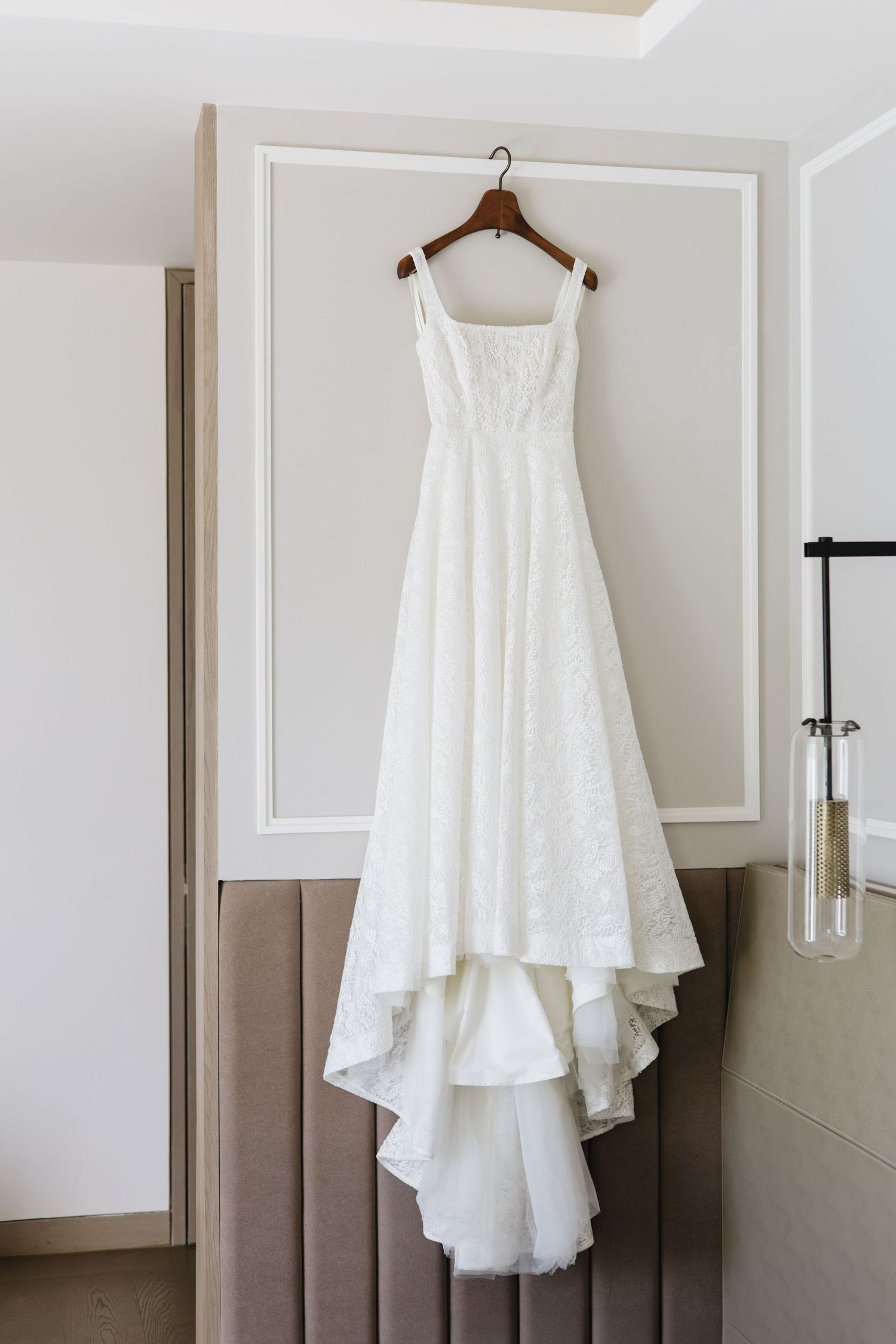 Elegant white wedding dress with lace detail and dramatic train hangs on wooden hanger against neutral wall backdrop.