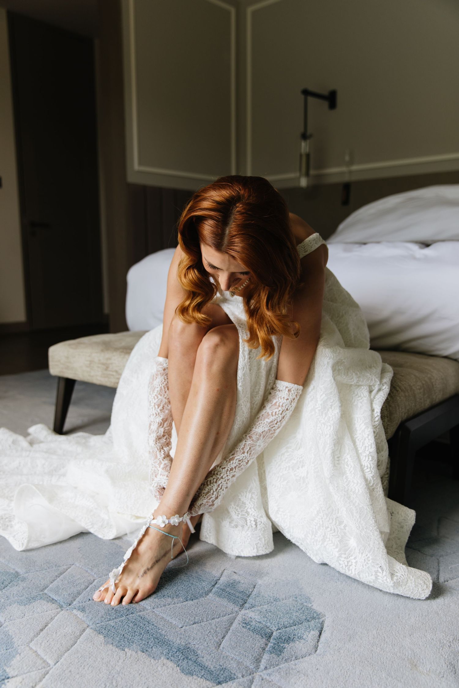 Elegant white lace dress and delicate anklet accessory shown on figure sitting on edge of bed in natural light.